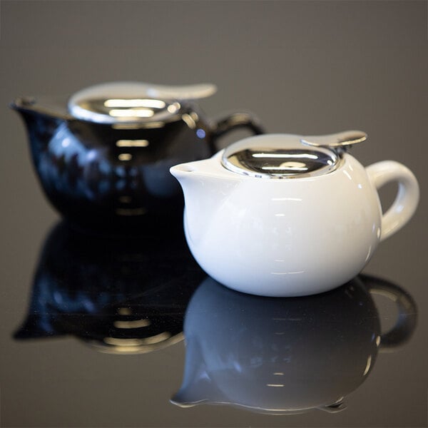 Two round ceramic teapots, one white and one black, with shiny metallic lids are placed on a reflective surface.