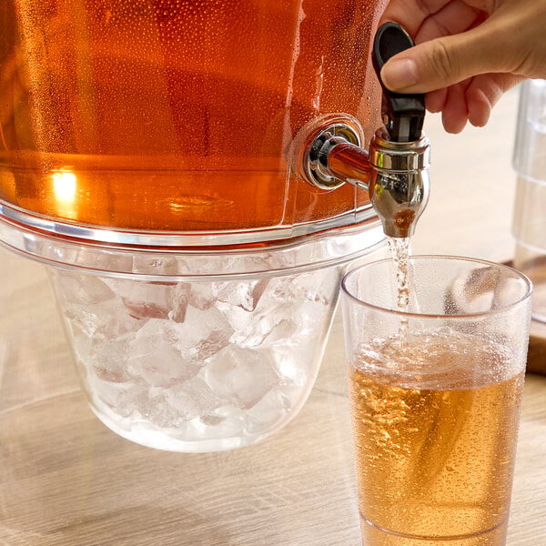 A hand is dispensing a cold amber-colored beverage from a clear container with an ice chamber into a glass.