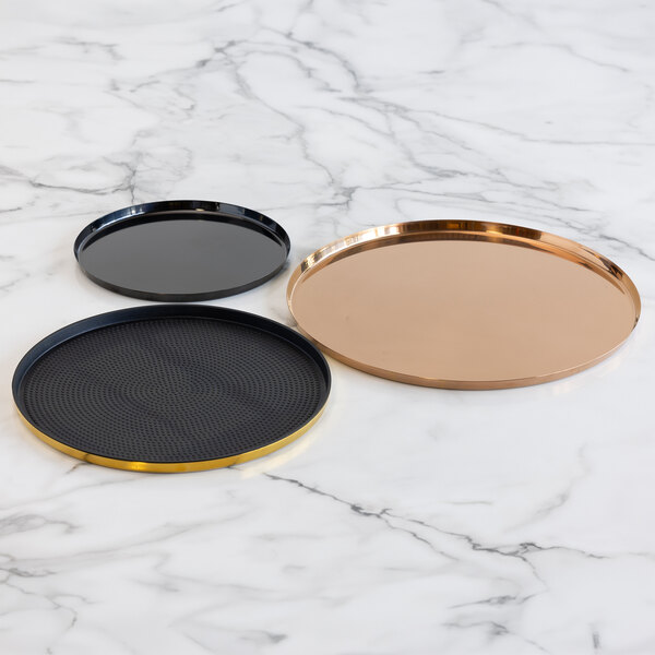 Three round serving trays of different sizes and colors are placed on a white marble surface; one is black with a gold rim and a textured non-slip insert, one is solid black with a glossy finish, and one is metallic gold or copper.