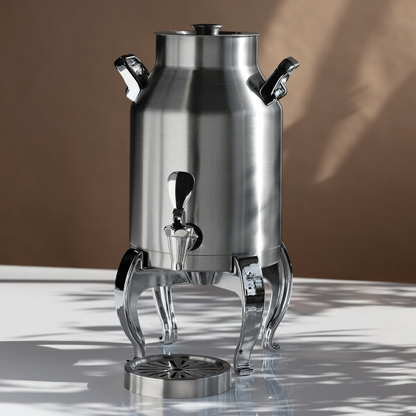 A stainless steel beverage urn with a spigot, two side handles, and four curved legs, placed on a reflective surface with a round drip tray in front.