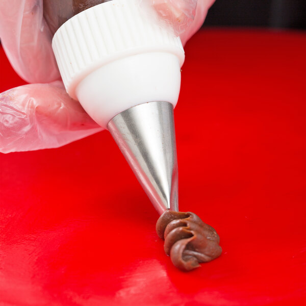 A close-up of a gloved hand using a piping bag with a metal tip to pipe chocolate frosting onto a red surface.
