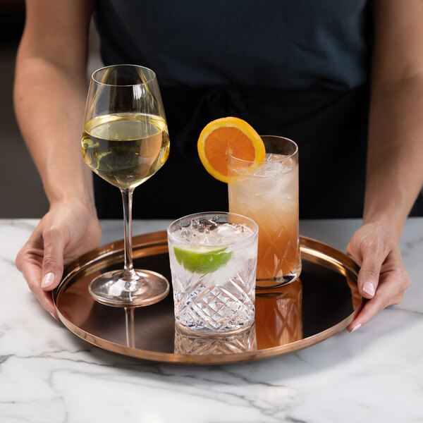 A person is holding a round rose gold tray with three different drinks: a glass of white wine, a cocktail with a lime wedge, and a tall drink garnished with an orange slice.