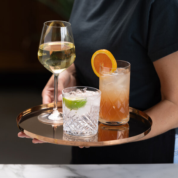 A person is holding a round, rose gold tray with three different drinks: a wine glass with white wine, a short glass with a lime wedge and ice, and a tall glass with an orange slice and a mixed drink.