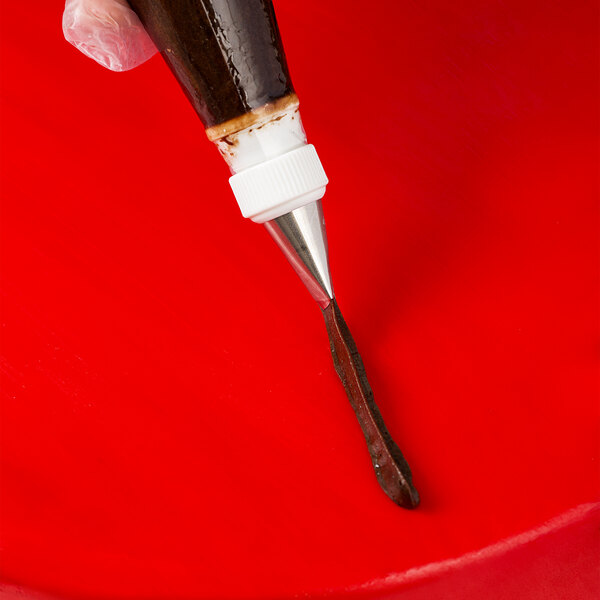 A close-up of a piping bag with a metal ruffle tip dispensing chocolate icing onto a bright red surface.