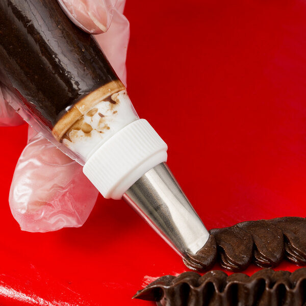 A close-up of a gloved hand using a piping bag with a metal ruffle tip to pipe chocolate frosting onto a red surface.