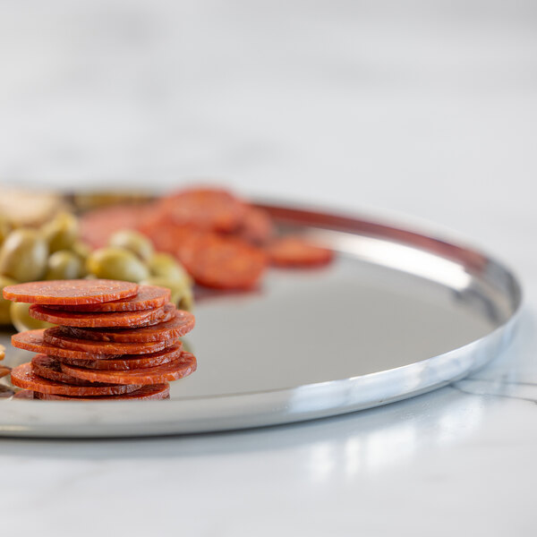 A round, polished metal serving tray with a stack of sliced pepperoni and some green olives on it.