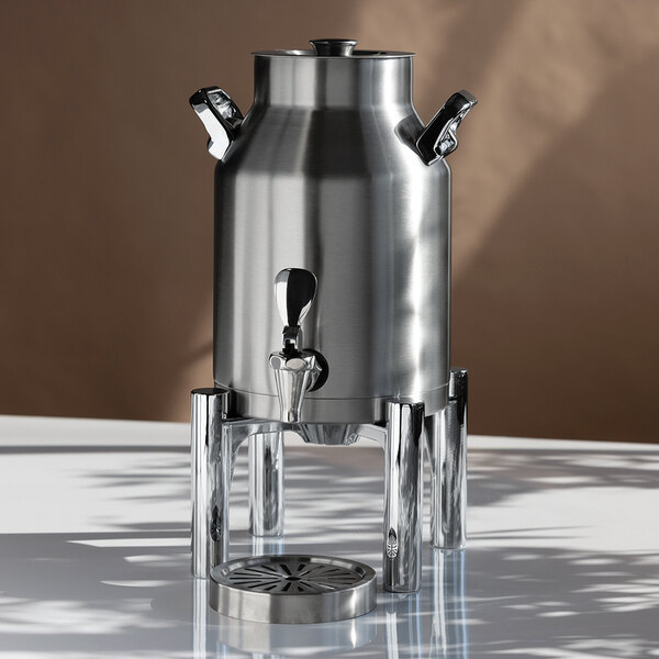 A stainless steel beverage urn with a spigot, handles, and a stand, placed on a reflective surface.