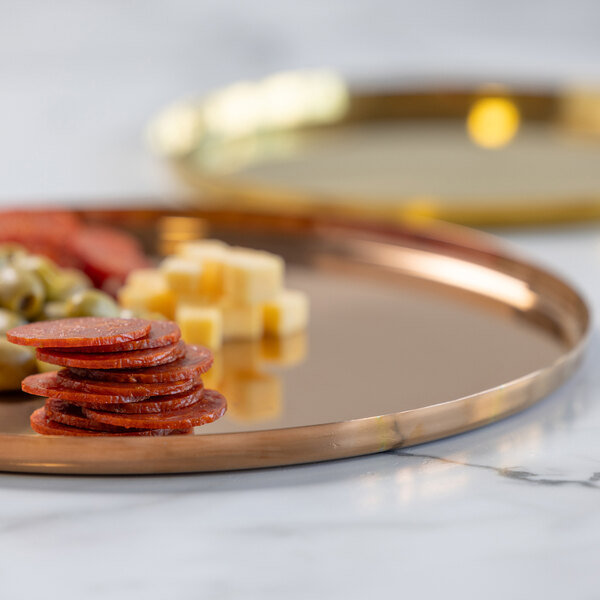 A round rose gold tray with a reflective surface, holding sliced cured meat, cubed cheese, and green olives.