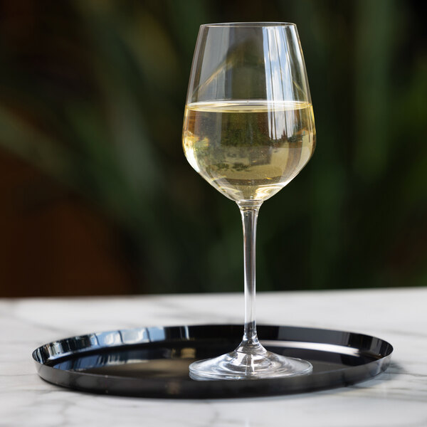 A glass of white wine sits on a round black serving tray placed on a marble surface.
