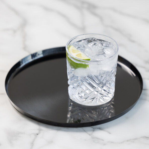 A clear glass with a textured pattern, filled with ice, a lime wedge, and a clear beverage, sits on a round black tray atop a marble surface.