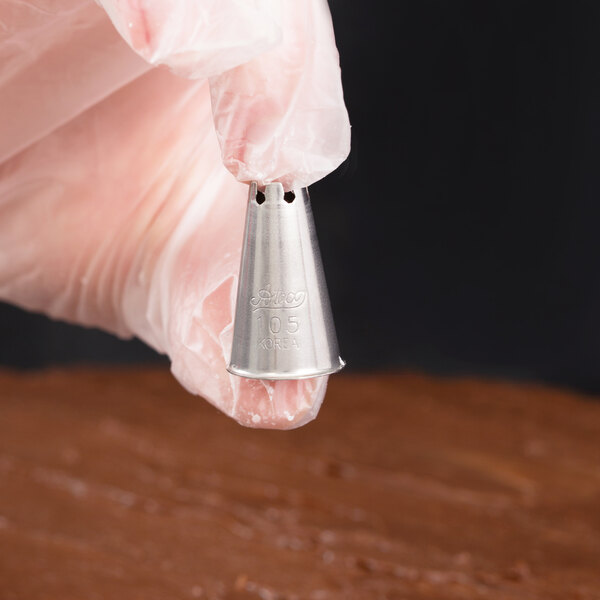 A gloved hand is holding a small metal piping tip with the number 105 engraved on it, above a chocolate-frosted surface.