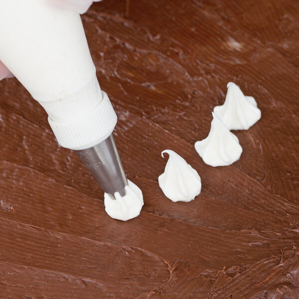 A piping bag fitted with a metal tip is being used to pipe white frosting in decorative shapes onto a chocolate-frosted surface.