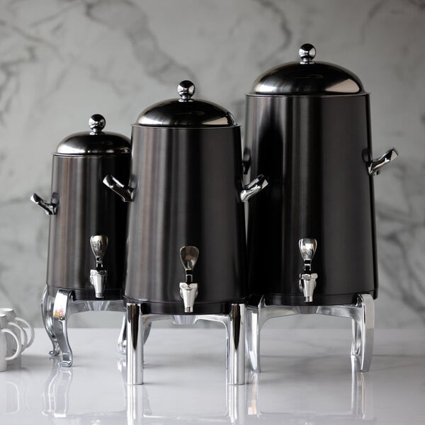 Three black and silver stainless steel beverage urns with domed lids and spigots are displayed on a marble surface.