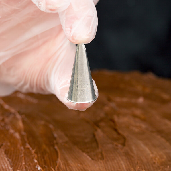 A gloved hand is holding a metal piping tip above a surface covered with chocolate frosting.