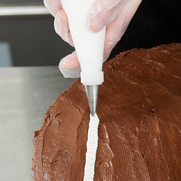 A hand wearing a glove is using a piping bag with a metal tip to pipe a white decorative line onto a chocolate-frosted cake.