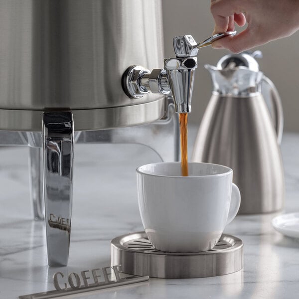 A hand is dispensing coffee from a stainless steel urn into a white cup placed on a metal coaster, with another stainless steel carafe in the background.