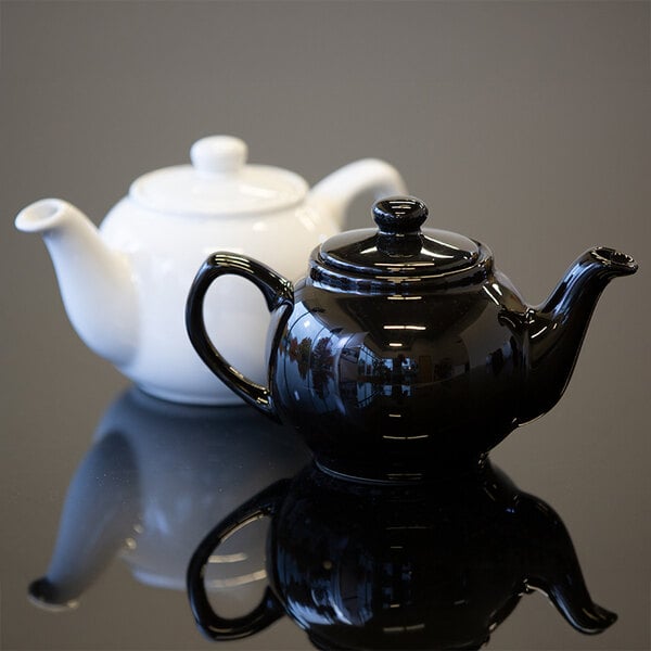 Two ceramic teapots, one black and one white, are placed on a reflective surface.
