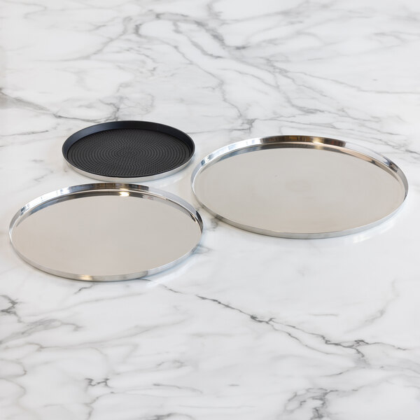Three round serving trays are displayed on a marble surface, two with a polished stainless steel finish and one with a black non-slip insert.