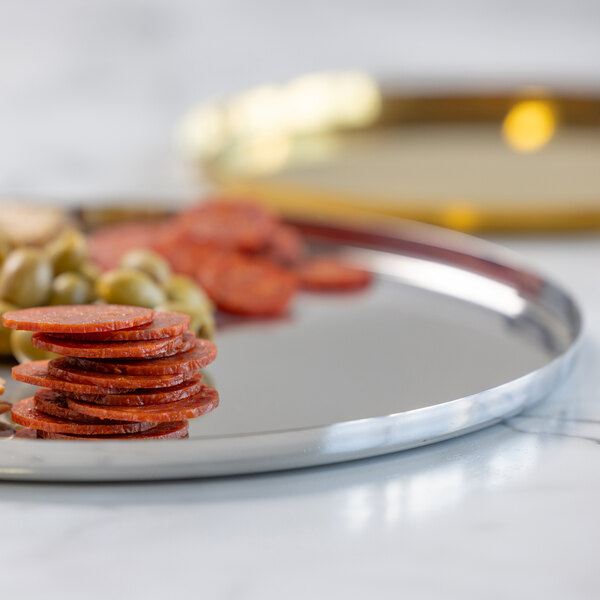 A round, polished stainless steel serving tray with a non-slip insert, holding sliced cured meat and green olives.