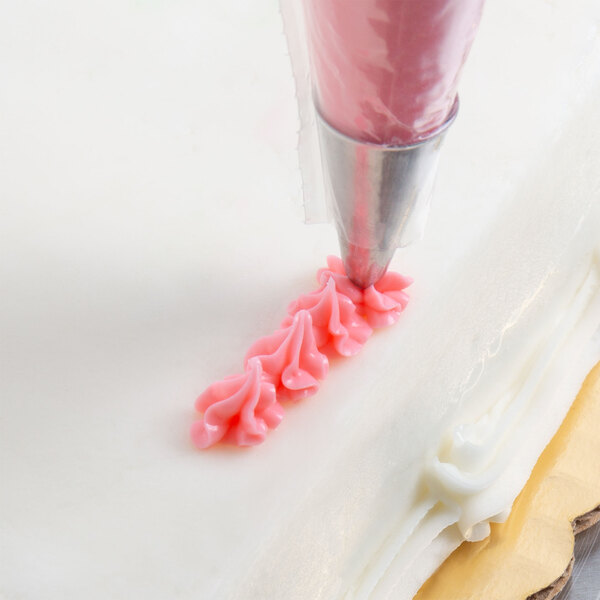 A close-up of a piping bag with a metal tip applying pink icing in a decorative pattern on a white frosted cake.