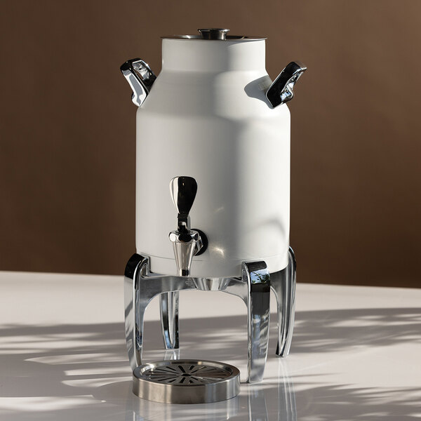 A white, cylindrical beverage urn with chrome handles and spout, elevated on a shiny metal stand with a drip tray beneath.