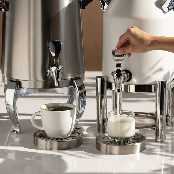 A hand is dispensing milk from a white and stainless steel beverage urn into a glass, next to another urn with a cup of coffee underneath its spout.