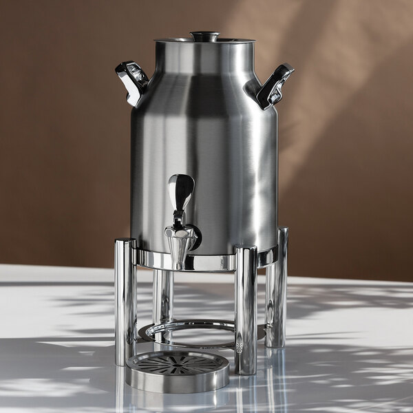 A stainless steel beverage urn with a spigot, two side handles, and a stand, placed on a reflective surface.