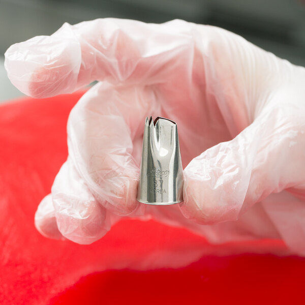 A gloved hand is holding a metal piping tip with a jagged opening, commonly used for cake decorating.