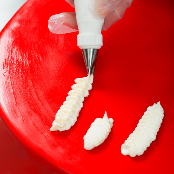 A gloved hand is using a piping bag with a metal tip to pipe white icing in a ruffled pattern onto a red surface.
