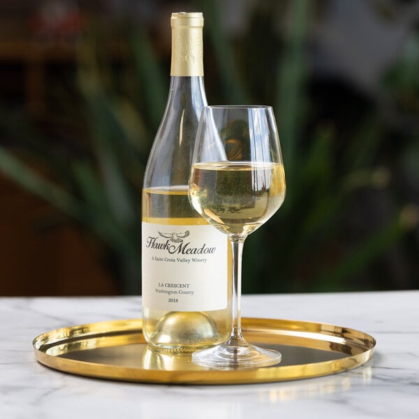 A bottle of white wine and a filled wine glass are placed on a round, gold-colored serving tray atop a marble surface.