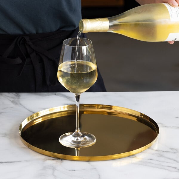 A glass of white wine is being poured from a bottle, standing on a round, gold-colored serving tray placed on a marble surface.
