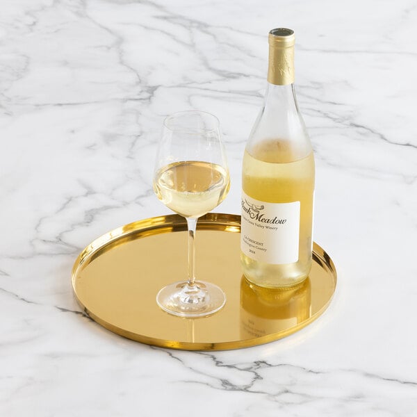 A gold round serving tray holds a bottle of white wine and a filled wine glass on a marble surface.