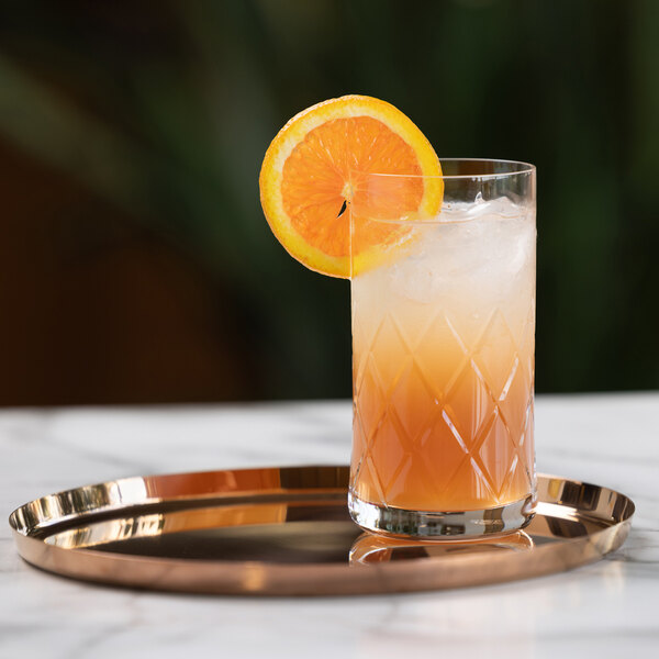 A tall glass filled with an orange and white layered drink, garnished with an orange slice, sits on a round rose gold tray on a marble surface.