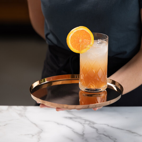 A person is holding a round, reflective rose gold tray with a tall glass of iced orange beverage garnished with an orange slice.
