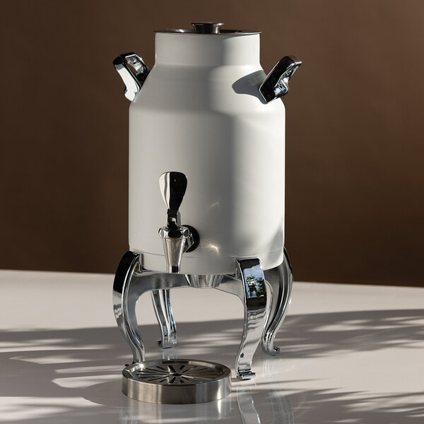 A white, cylindrical beverage urn with chrome handles and legs, featuring a spigot and a removable drip tray, placed on a reflective surface.