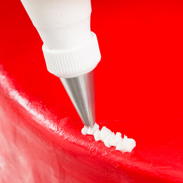 A close-up of a metal piping tip applying white icing to the edge of a red frosted cake.