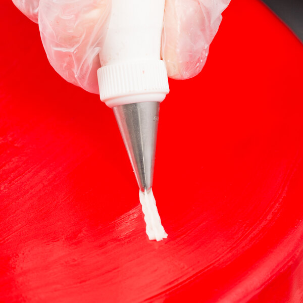 A close-up of a gloved hand using a metal piping tip to pipe white icing onto a red surface.