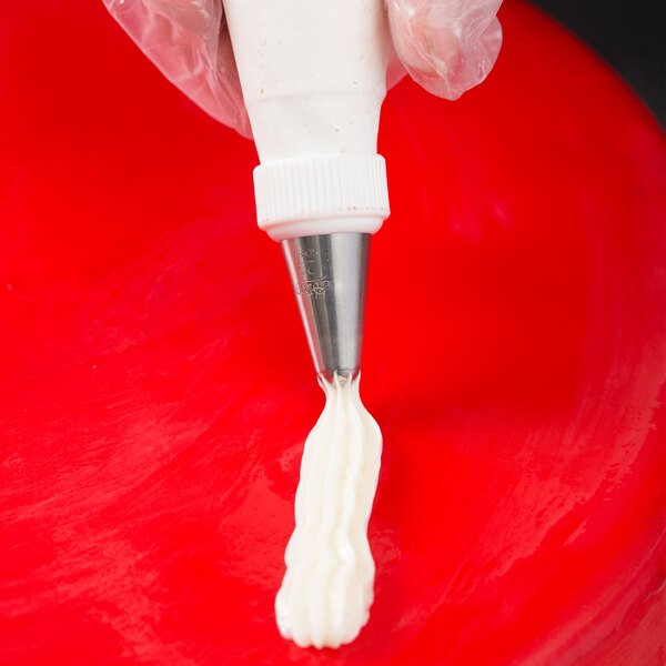 A close-up of a gloved hand using a piping bag with a metal star-shaped tip to pipe white icing onto a red surface.