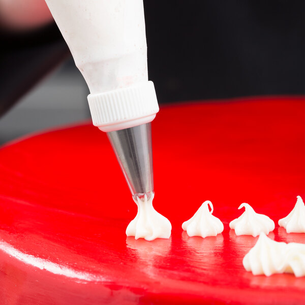 A close-up of a piping bag with a metal star tip applying white icing onto a smooth red surface.