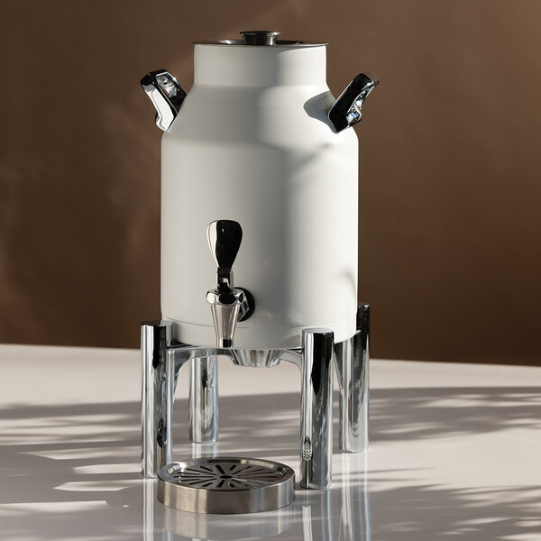 A white, cylindrical beverage urn with chrome handles and spigot, elevated on a shiny metal stand with a drip tray beneath.