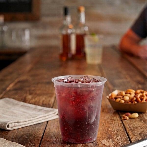 A clear plastic cup filled with a red iced beverage sits on a rustic wooden table next to a napkin and a bowl of mixed nuts.