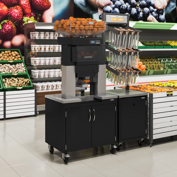 A commercial juicing machine with a basket of oranges on top, positioned on a black cabinet with wheels in a grocery store produce section.