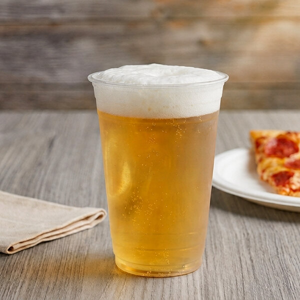 A clear plastic cup filled with a light, foamy beverage sits on a wooden table next to a folded napkin and a plate with a slice of pizza.
