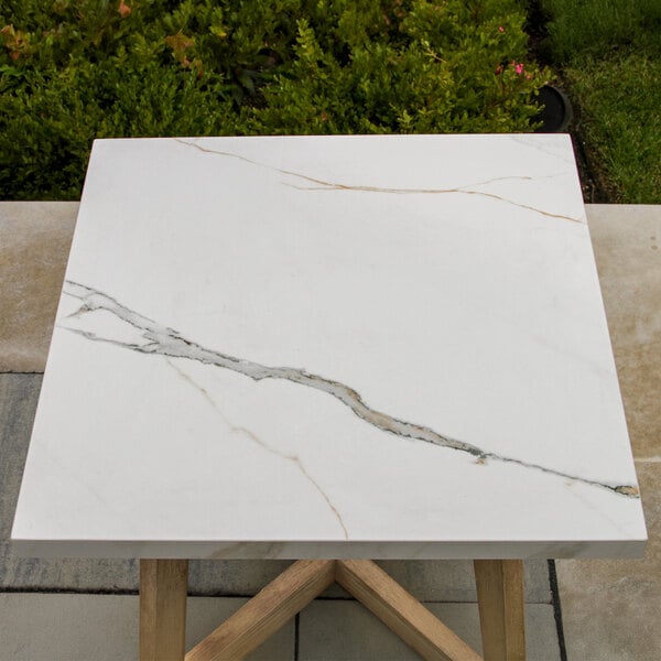 A square table with a white marble-like top featuring gray and tan veining, supported by light wood legs.