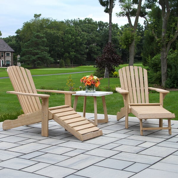 Two wooden Adirondack chairs with matching footrests are positioned on a stone patio, flanking a small square table with a white top that holds a vase of orange flowers, set against a backdrop of green grass and trees.