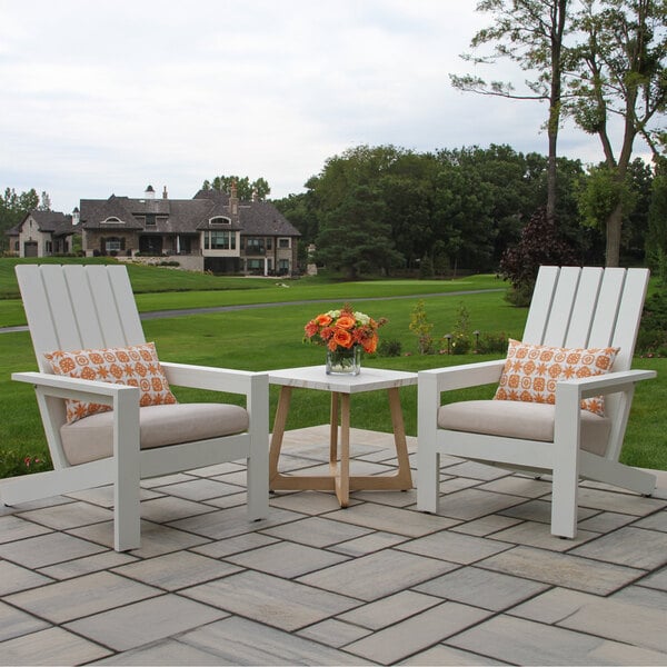 Two white wooden chairs with beige cushions and patterned pillows are positioned on a stone patio, with a small square table between them holding a vase of orange flowers, set against a backdrop of green lawn and trees.