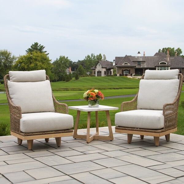 Two cushioned wicker chairs are placed on a patio with a small square table between them, topped with a vase of flowers, and a large house and green lawn are visible in the background.