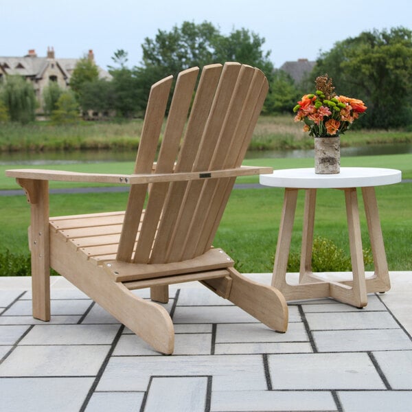 A wooden Adirondack chair with a built-in ottoman is placed on a patio next to a small round side table with a vase of orange flowers, overlooking a grassy area and pond.
