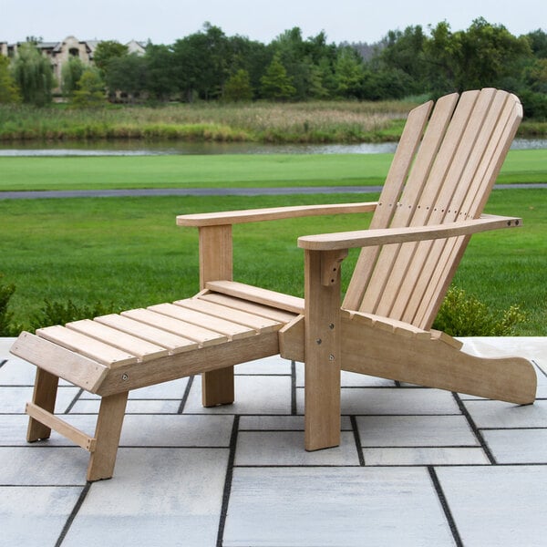 A wooden Adirondack chair with a built-in ottoman is placed on a tiled patio overlooking a grassy area and a pond.
