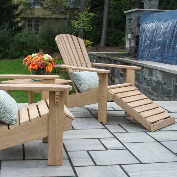 Two wooden Adirondack chairs with slatted backs and seats, one featuring a built-in ottoman, are placed on a stone patio next to a small table with a flower arrangement.